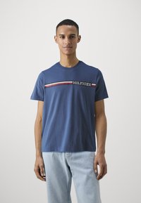 Modrá bavlnená tričko má okrúhly výstrih a horizontálne pruhované detaily s textom "HILFIGER" v kontrastných farbách.