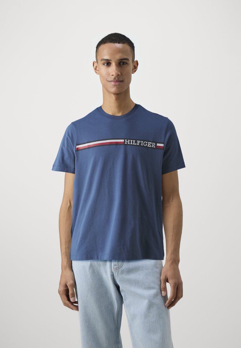 Modrá bavlnená tričko má okrúhly výstrih a horizontálne pruhované detaily s textom "HILFIGER" v kontrastných farbách.