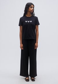 Μαύρο βαμβακερό T-shirt με τρία σχέδια καρδιών σε σειρά στο μπροστινό μέρος. Φορεμένο με μαύρο παντελόνι φαρδιά μπατζάκια. Το μοντέλο στέκεται με τα χέρια στα πλάγια.