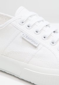 Bílé plátěné tenisky s bílými tkaničkami, stříbrnými očky a látkovou visačkou "Superga" na boku poblíž podrážky.