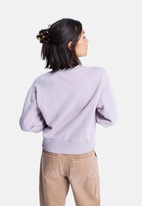 Felpa lilla con vestibilità rilassata; polsini e orlo a coste; texture morbida. Indossata con pantaloni beige; capelli raccolti con una clip scura a forma di fiore.
