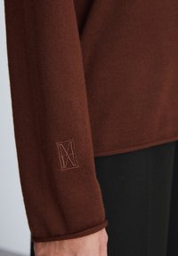 Brązowy sweter dzianinowy z rękawem, na którym znajduje się subtelnie wyhaftowane logo na mankiecie. Gładka tekstura, luźny krój oraz wywinięty brzeg.