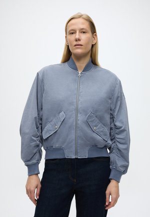 Femme aux cheveux blonds raides portant une veste bomber bleu clair à fermeture éclair avec des poches à rabat et un jean bleu foncé, debout devant un fond uni.