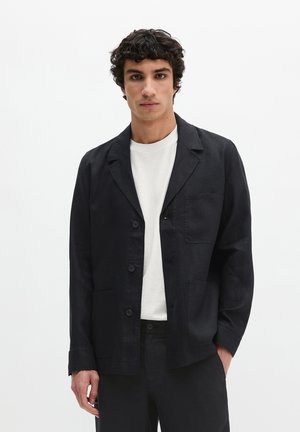 Männliches Model trägt schwarzen, geknöpften Casual-Blazer über weißem Hemd und steht vor weißem Hintergrund.