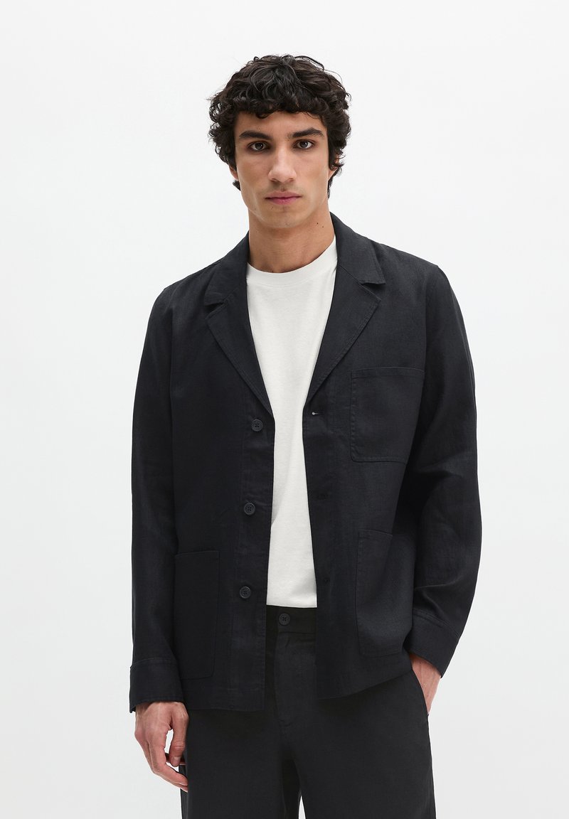 Male model wearing black buttoned casual blazer over white shirt, standing against plain white background.