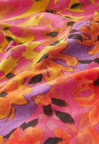Tissu floral présentant des teintes vives de rose, jaune et violet avec une surface texturée. Des formes de pétales et des accents noirs sont visibles.