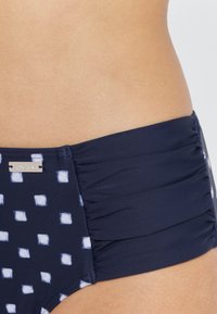 Marineblaue Badehose mit weißen quadratischen Mustern, mit gerafften Seiten und einem silbernen Markenzeichen. Glatte Textur, elastisches Material.