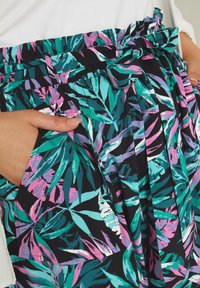 Kukkakuvioinen hame mustassa kankaassa, jossa on monivärisiä trooppisia lehtikuvioita, laskostettu muotoilu ja sidottava vyötärö, jossa on sivutaskut.