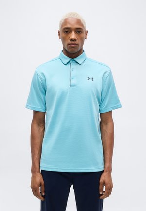 Homme aux cheveux blonds courts portant un polo Under Armour bleu clair et un pantalon foncé, debout devant un fond blanc uni.