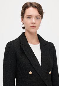 Juodas, tekstūruotas dvigubas švarkas su auksinės spalvos mygtukais ir plačiomis apykaklėmis, uždėtas ant balto megztinio.