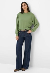 Pullover verde a maniche lunghe, vestibilità rilassata, abbinato a jeans larghi in denim scuro a vita alta. Cintura con stampa animale, scarpe a punta.