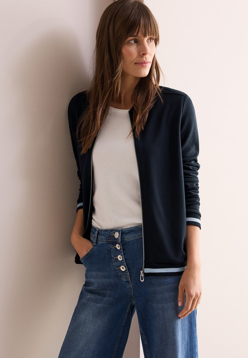 Marineblå zip-jacka med vita randiga muddar, kombinerad med ljusgrå t-shirt och högmidjade denimjeans med knapprad.
