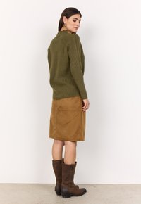 Olivgrüner gerippter Pullover kombiniert mit einem braunen Wildlederrock in Knielänge und braunen ledernen Stiefeln bis zum Knie mit niedrigem Blockabsatz.