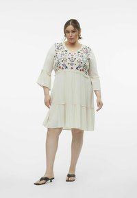 Vestido blanco con bordado floral en varios colores en el pecho, mangas acampanadas y diseño de falda en capas. Lleva sandalias negras.