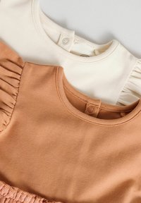 Deux t-shirts pour enfants : un pêche avec une texture lisse et un col rond, et un crème avec des manches côtelées et une fermeture à boutons à l'arrière.