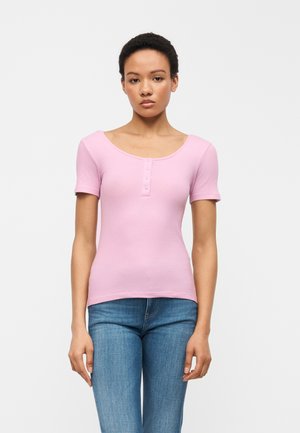 Νεαρή γυναίκα στέκεται με το πρόσωπο προς τα εμπρός, φορώντας ένα ροζ ανοιχτόχρωμο ριμπ κοντομάνικο τοπ με κουμπιά και μπλε τζιν, με φόντο μια απλή λευκή επιφάνεια.