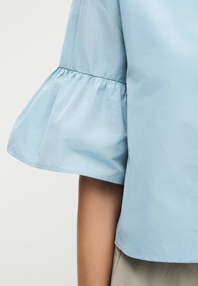 Haut bleu clair à manches courtes avec un détail froncé évasé au-dessus du coude sur le bras d'une personne, sur un fond uni.