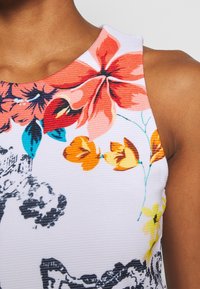 Gros plan sur un vêtement sans manches blanc à texture côtelée, décoré de grands motifs floraux rouges, orange, jaunes et noirs près de l'épaule.