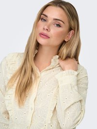 Licht crèmekleurige blouse van gestructureerde stof, met bloemrijke borduursels, geplooide accenten langs de voorkant en mouwen, en knoopsluitingen.
