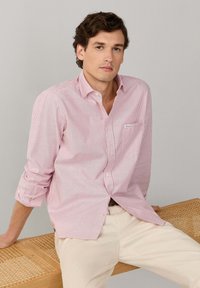 Chemise à boutons rayée rouge et blanc avec une poche poitrine, manches retroussées et texture en coton. Le modèle porte un pantalon beige clair.