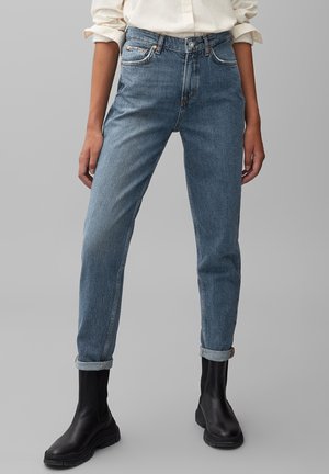 Osoba ubraną w podwinięte niebieskie dżinsy, kremową koszulę z podwiniętymi rękawami i czarne masywne botki na jednolitym szarym tle.