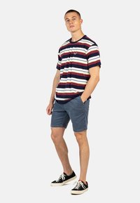 Camiseta de manga corta a rayas que presenta rayas horizontales en rojo, azul marino y blanco; se lleva con pantalones cortos grises y zapatillas negras.