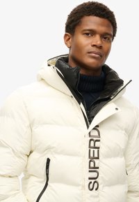 Puffet jakke i creme med en sort inderside, featuring vandret quiltning, lynlås lommer og et fremtrædende logo på fronten.