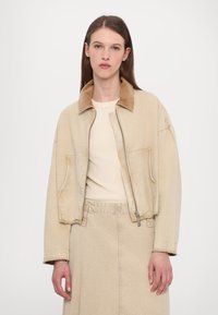 Giacca in denim beige con colletto in corduroy marrone a contrasto, chiusura con zip, due tasche frontali e vestibilità rilassata. Indossata sopra un top color crema.