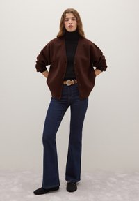 Brauner, übergroßer Cardigan mit Knöpfen, getragen über einem schwarzen Rollkragenpullover. Kombiniert mit ausgestellten dunklen Jeans und schwarzen Slip-On-Schuhen.