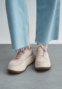Baskets plateforme en suède beige avec bouts ronds, devants lacés et détails texturés, associées à un jean bleu clair à ourlet effrangé.