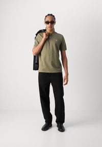 Tricou polo verde măslin cu mâneci scurte, pantaloni negri și pantofi negri. Jachetă din piele aruncată pe umăr, ochelari de soare purtați.