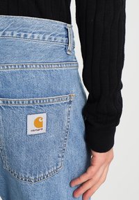 Jasnoniebieskie dżinsy o wysokim stanie, z detalicznym przeszyciem i tylną kieszenią z białą naszywką z żółtym symbolem.