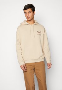 Felpa beige con cappuccio, cordini, tasca frontale e grafica di una farfalla marrone con il testo: "L'abbondanza è una mentalità." Indossata con pantaloni marroni chiari.
