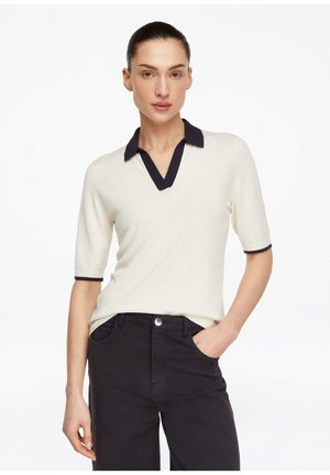 Kobieta stoi z jedną ręką w kieszeni, ubrana w kremową koszulkę polo z krótkim rękawem, czarnym kołnierzykiem i czarne spodnie na białym tle.