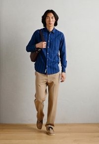 Cămașă din denim cu nasturi, de culoare bleumarin, cu dungi verticale, pantaloni bej și pantofi maronii. Poartă o geantă de umăr maronie. Podea din lemn deschis.