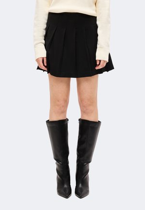 Mujer con falda negra plisada, suéter crema de manga larga y botas de cuero negras hasta la rodilla, de pie frente a un fondo liso.
