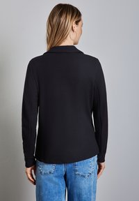 Camicia nera, a trama, con colletto e maniche lunghe, abbinata a jeans chiari a vita alta, vista da dietro.