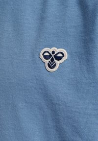 Jasnoniebieska bawełniana koszulka z haftowanym białym i granatowym logo, przedstawiającym stylizowany wzór owada po lewej stronie.