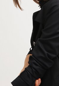 Abrigo negro de trench con cierre de un solo botón, cuello estructurado y mangas enrolladas con un acento en beige contrastante en el puño.