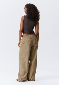 Tumši brūns bezroku tops, vaļīgas gaismas brūnas bikses ar elastīgu jostasvietu un sānu kabatām, gluda tekstūra, modelis redzams no muguras.