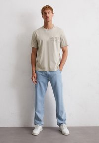 Light beige short-sleeve T-shirt with printed text on the chest, paired with light blue straight-leg jeans and white sneakers.