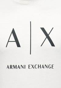Λευκό ύφασμα με μαύρο λογότυπο "A | X" πάνω από το κείμενο "ARMANI EXCHANGE" τυπωμένο σε αυτό.