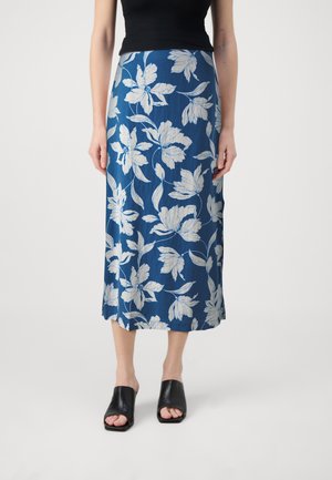 Femme portant une jupe midi bleue à imprimé floral blanc, un haut noir sans manches et des mules à talons noires à bout ouvert, debout devant un fond uni.