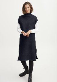 Dunkelblaues Strickkleid mit hohem Ausschnitt, seitlichen Schlitzen und kurzen Ärmeln; überezogen mit einem weißen Langarmshirt; getragen mit schwarzen Knöchelstiefeln.