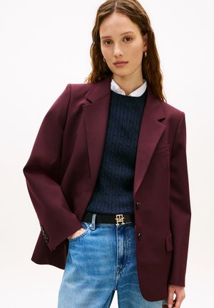 Vrouw die een bordeauxrode blazer draagt over een marineblauwe kabelgebreide trui, een wit overhemd met kraag, een blauwe spijkerbroek en een zwarte riem met een gouden gesp.