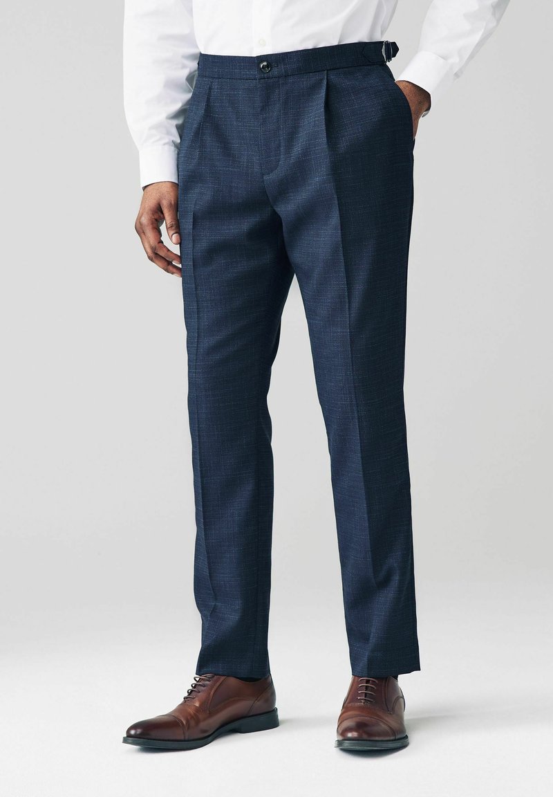 Pantalones de vestir de campana en color navy, con tejido texturizado y corte recto, que presentan presillas para el cinturón y un cierre de botón, combinados con zapatos de cuero marrón.