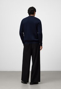 Marineblauer Pullover mit Rundhalsausschnitt, langen Ärmeln und glatter Textur, kombiniert mit schwarzen weiten Hosen und schwarzen Schuhen vor einem neutralen Hintergrund.
