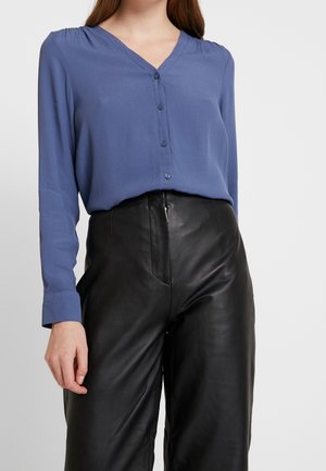 Femme portant un chemisier bleu à manches longues boutonné, rentré dans un pantalon en cuir noir taille haute, sur un fond uni.