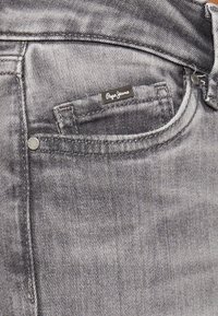 Jeans de denim gris con una textura suave. Entre sus características se incluyen un bolsillo delantero, etiqueta de marca y detalle de botón metálico.