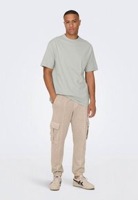 Tricou verde deschis cu mâneci scurte, asortat cu pantaloni cargo bej cu buzunare laterale, purtați cu adidași bej cu accente bleumarin.